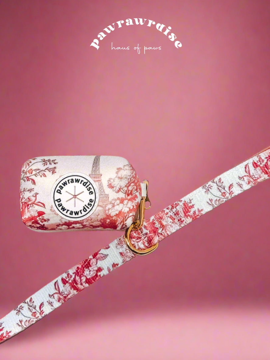 A neoprene poop bag holder with a floral pattern and a Pawrawrdise brand logo, featuring a dispense hole and a gold clasp attached on the leash, on a pink background - front view