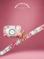 A neoprene poop bag holder with a floral pattern and a Pawrawrdise brand logo, featuring a dispense hole and a gold clasp attached on the leash, on a pink background - front view