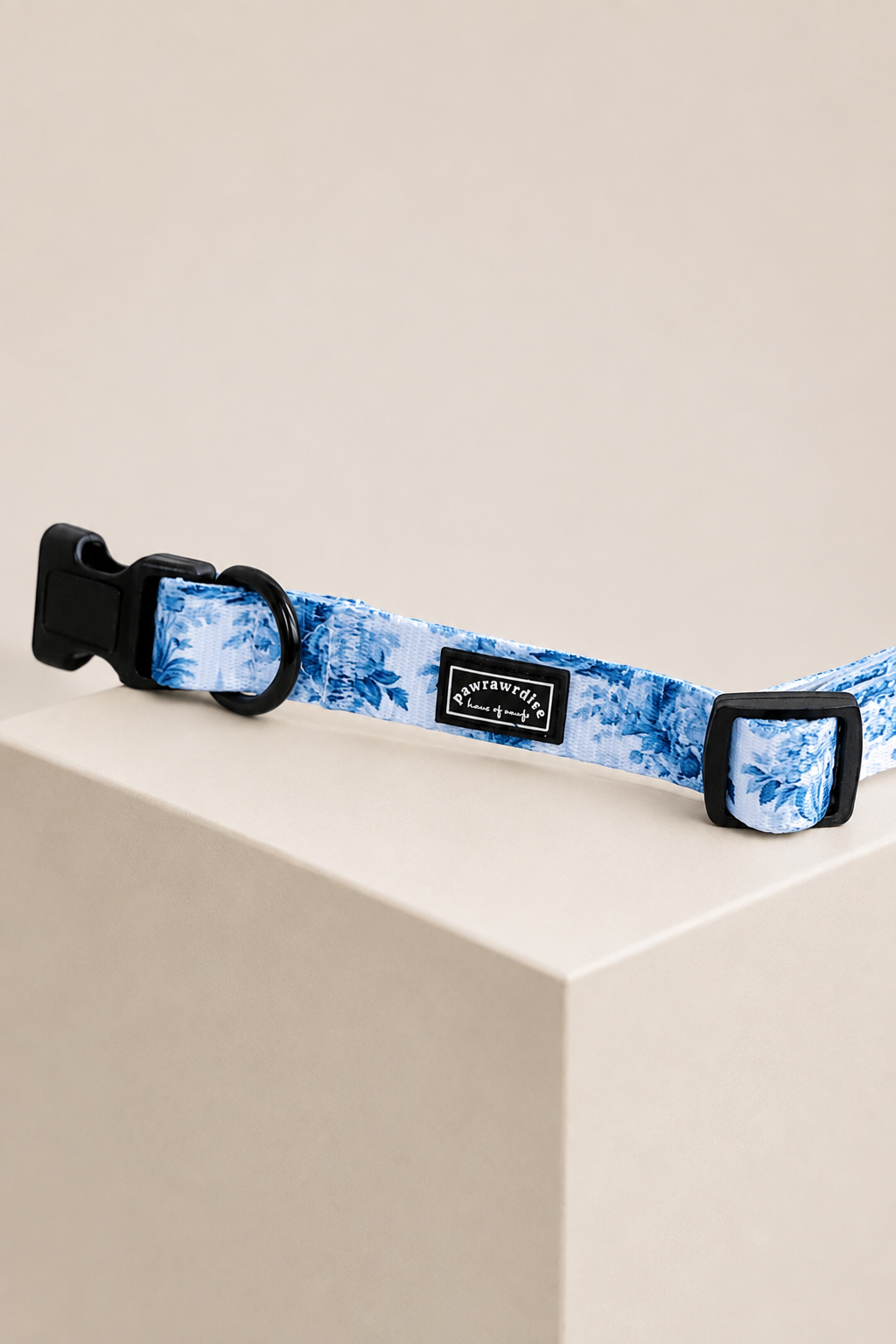 A close-up of Pawrawrdise Enchanted pet collar with a blue and white floral pattern, black plastic hardware, and a central black brand patch, resting on a beige display block.