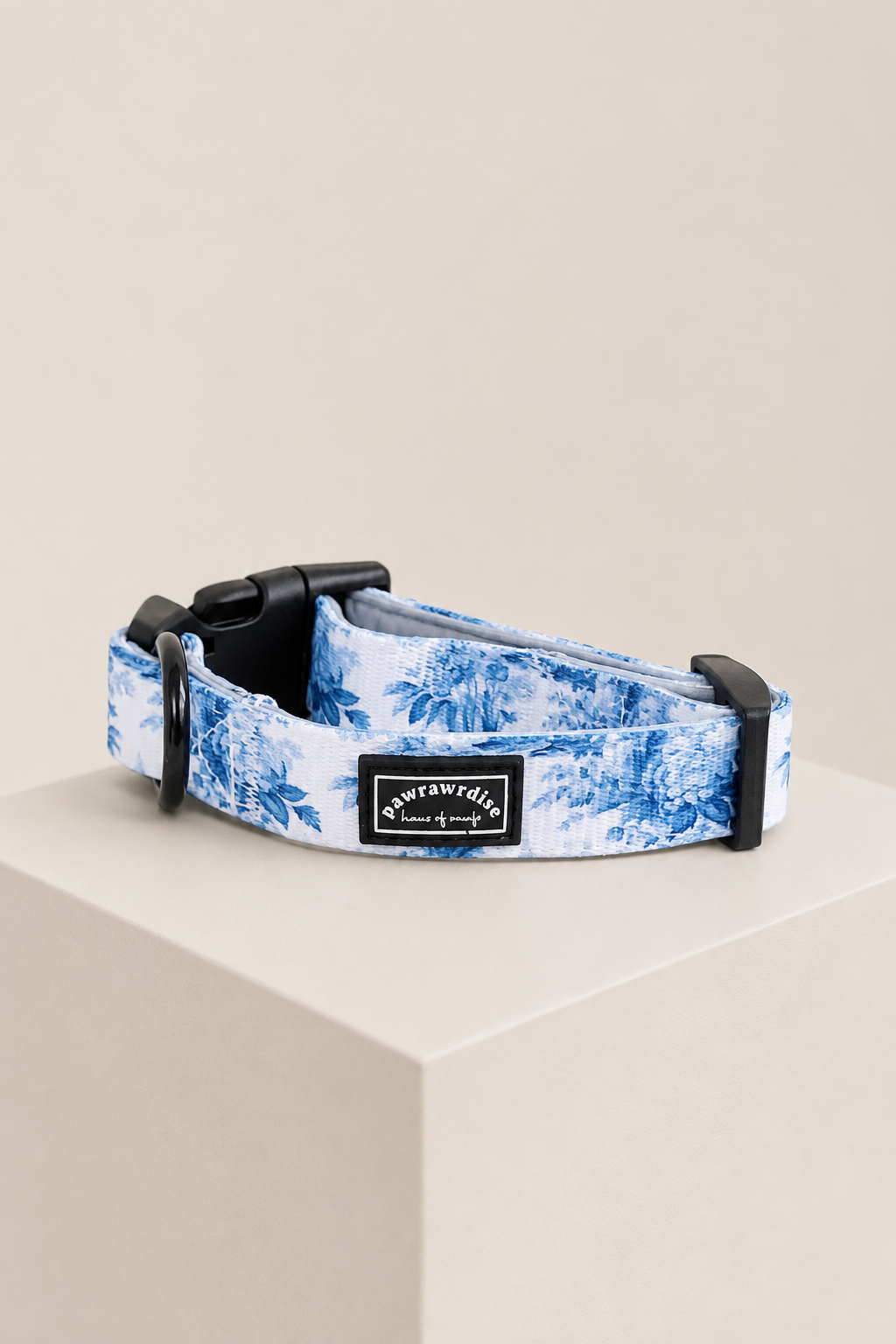 Pawrawrdise’s Enchanted blue and white floral patterned dog collar with black plastic hardware, displayed on a beige geometric pedestal against a neutral beige background. The brand label reads Pawrawrdise 