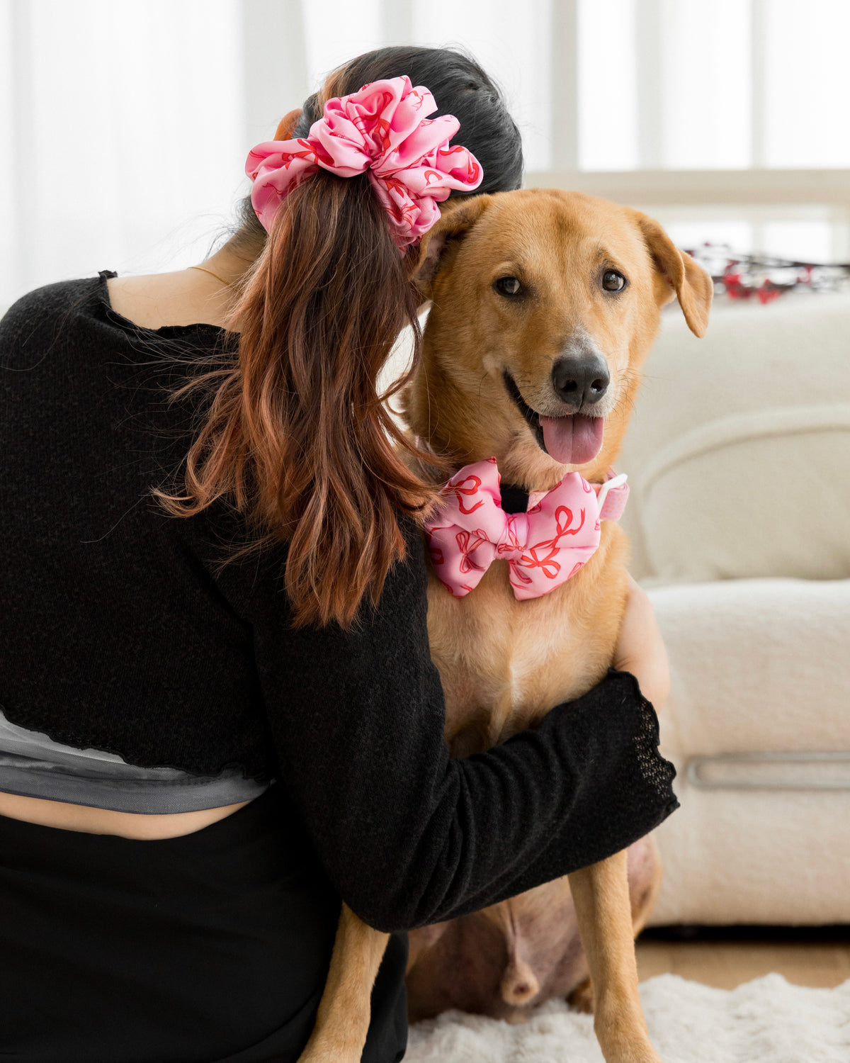 Human wearing a pink scrunchie and a dog wearing collar with a bow, both sitting on the floor.