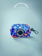 A multicolored batik print poop bag holder with a dispensing hole and a black clasp, placed against a blue background.
