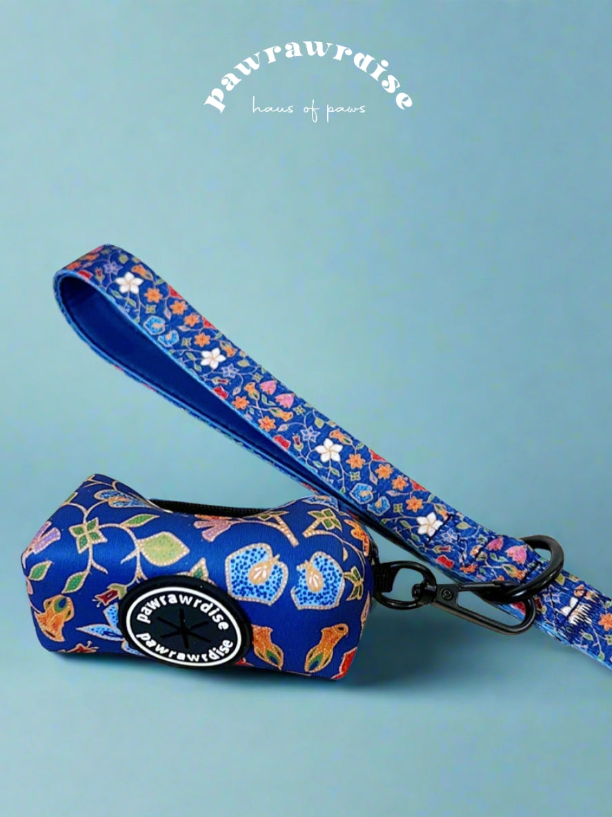 A multicolored batik print poop bag holder with a black clasp, placed against a blue background with leash