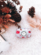 A neoprene poop bag holder with a Christmas-themed design, featuring a dispensing hole, placed on a white furry surface, surrounded by decorative pine cones and faux fur.
